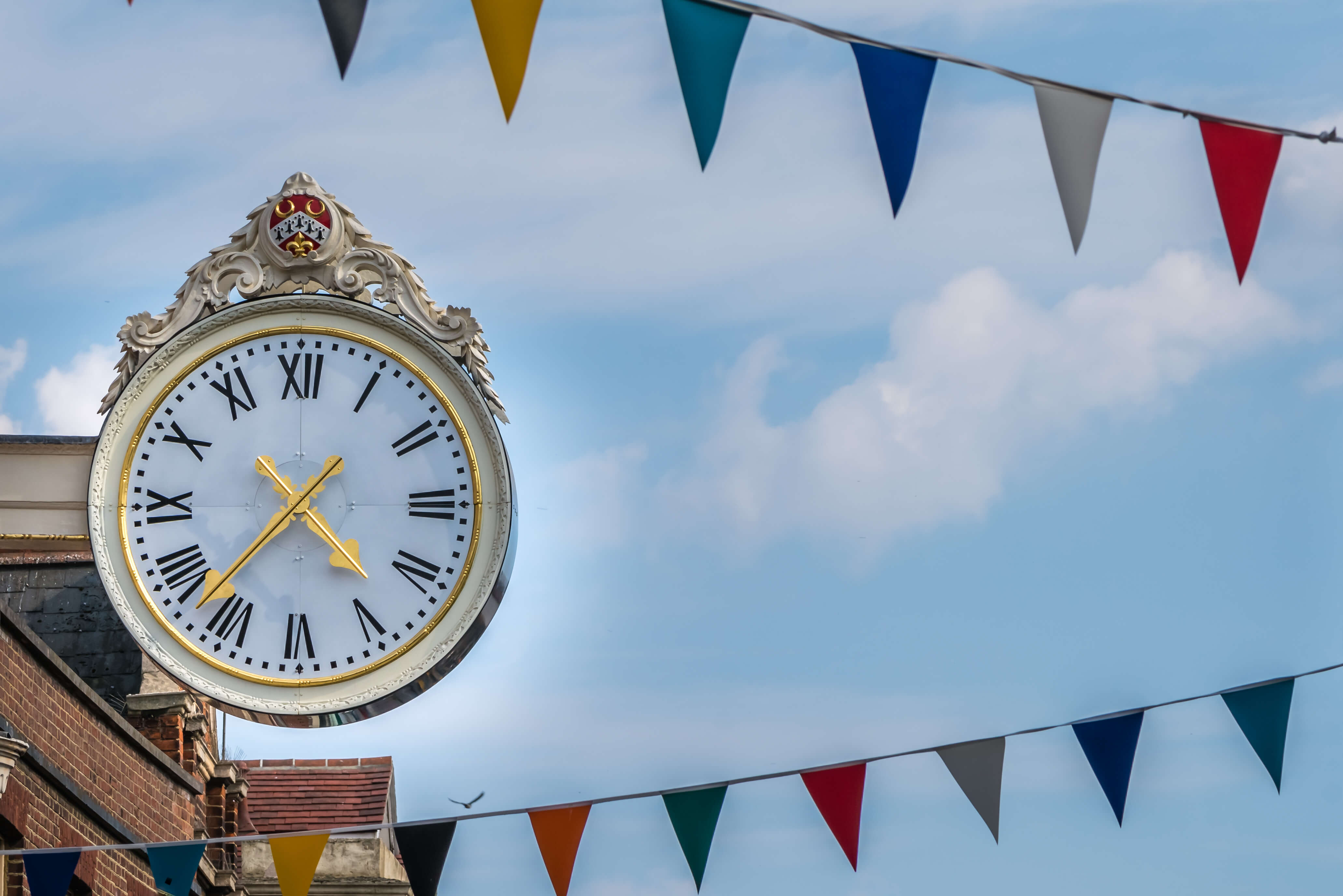 Ornate vintage theatre clock with Roman numerals and gold hands mounted on a historic building, with colourful bunting overhead—symbolising showtime and the anticipation of a live performance.